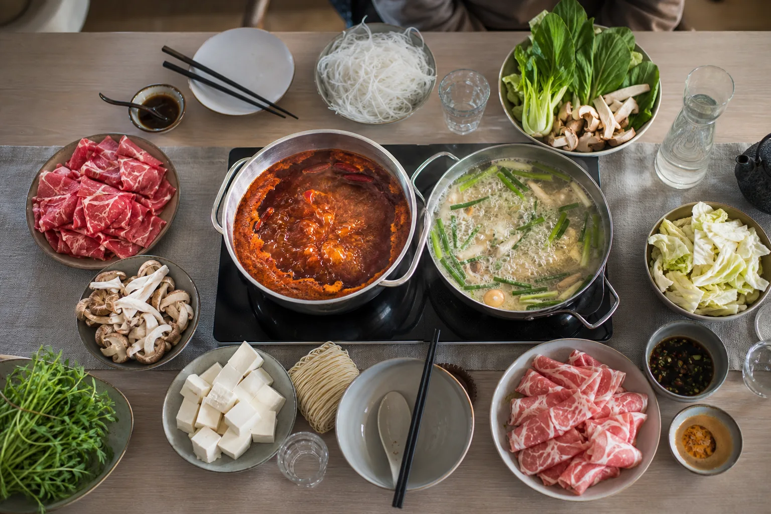 Ett sjudande hotpot-bord med två buljongskålar, tunt skivat kött, grönsaker och nudlar redo att doppas